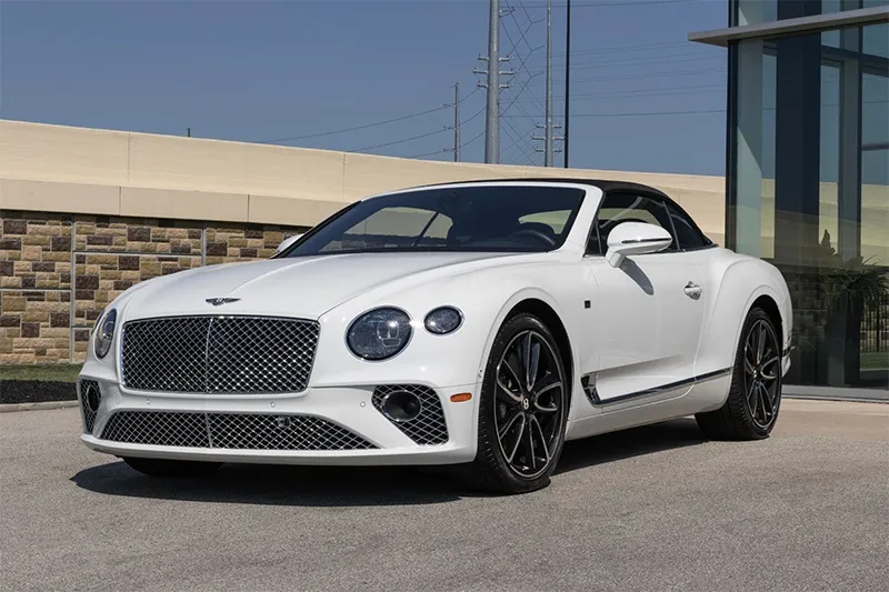 A white Bentley convertible sports car is parked outdoors on a paved surface near a building with glass walls, featuring black wheels and a sleek, modern design.