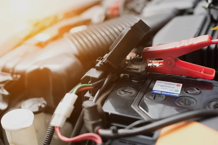 A close-up of a car battery with a red jumper cable clamp attached, surrounded by engine components under the hood. Sunlight is shining on the engine area.