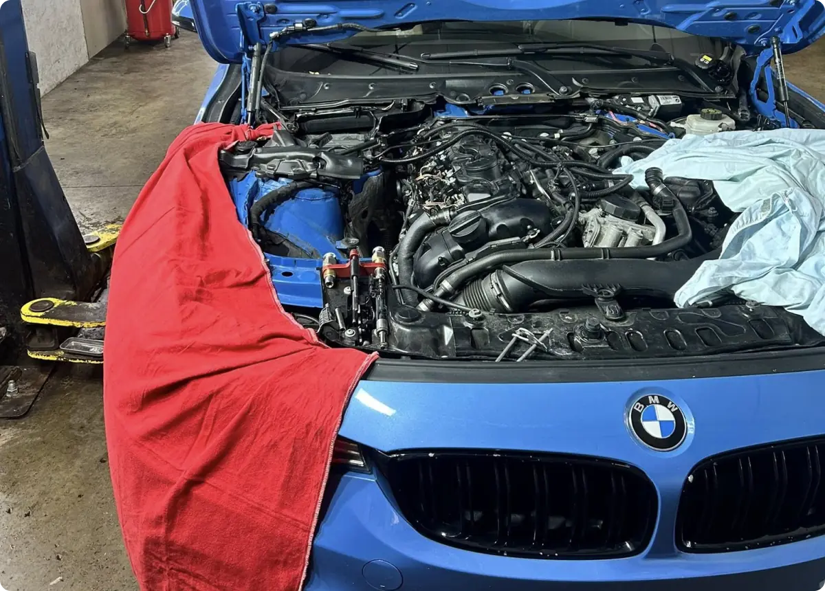 BMW repair in Bridgewater, MA at Holland’s Highline Automotive. A blue BMW car with its hood open is undergoing engine repairs in a garage. The front fenders are covered with red and light blue cloths to protect the paint. Various tools and engine components are visible.