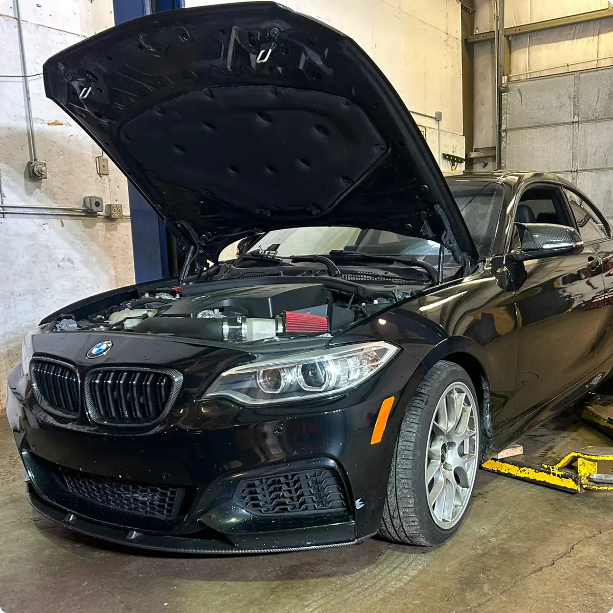 A black BMW car with its hood open is parked inside a garage or workshop. One front wheel is on a yellow lift. The car appears to be undergoing maintenance or repair.