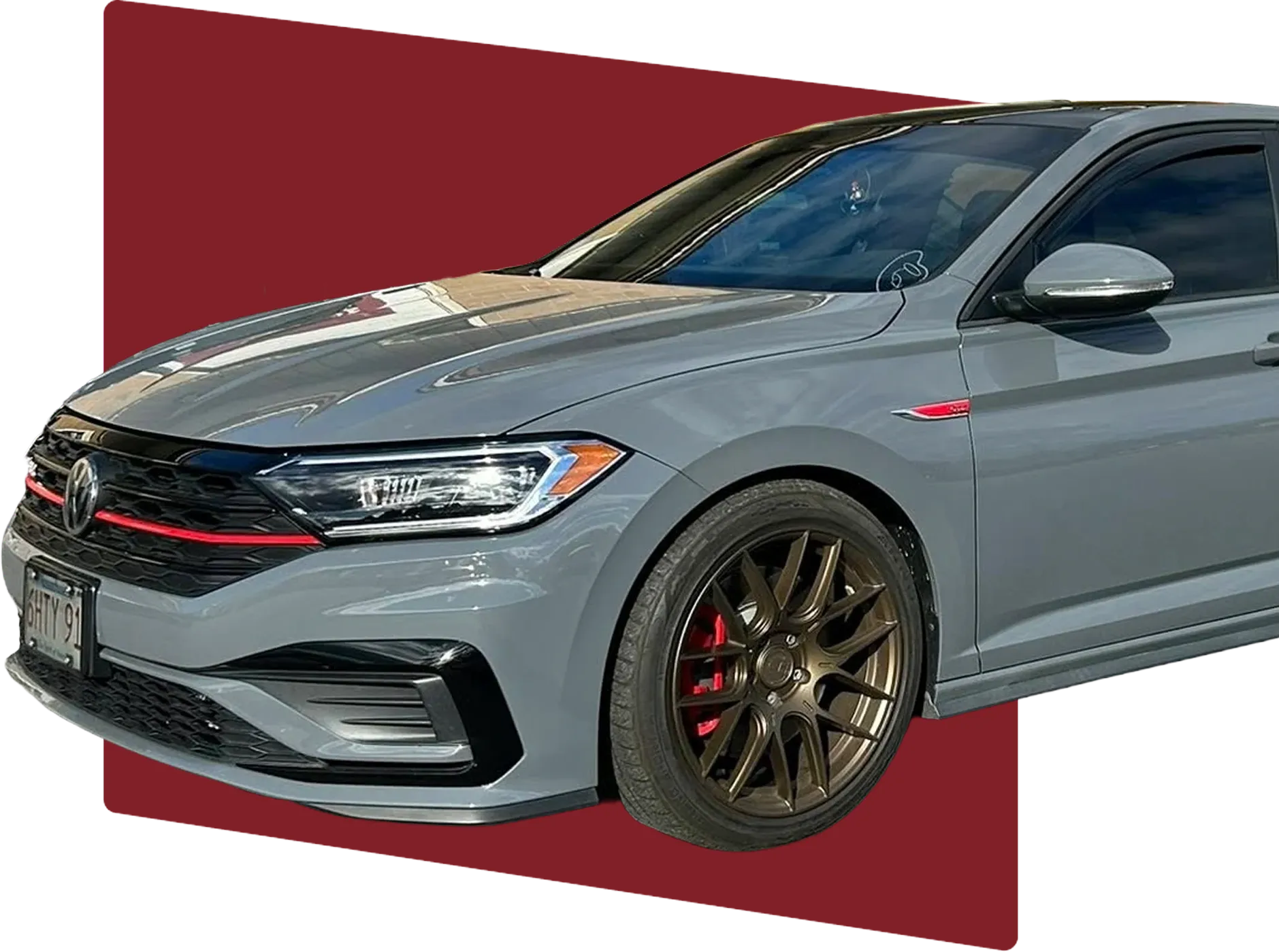 About us, auto repair in Bridgewater, MA at Holland’s Highline Automotive. A gray Volkswagen sedan with bronze aftermarket wheels and red brake calipers is parked, partially cropped on a maroon and pink background. The front and side of the car are visible.