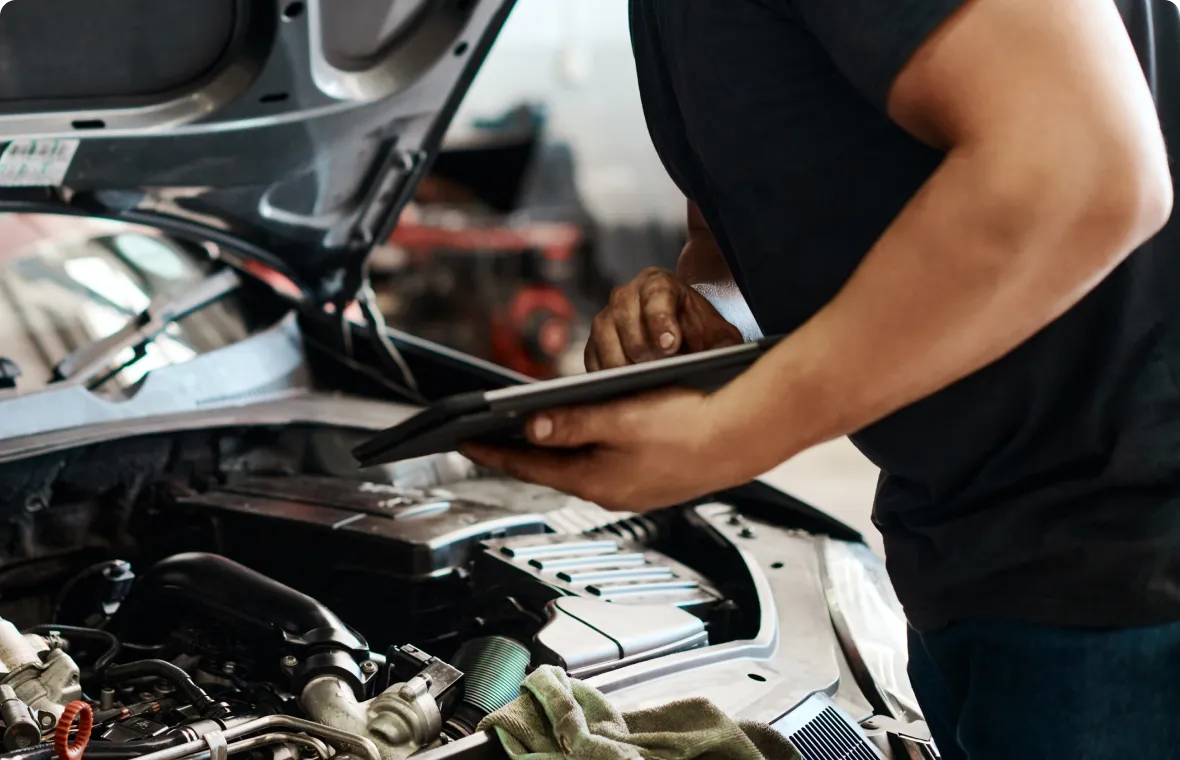 A person stands beside an open car hood, holding a tablet and inspecting the engine. Various engine components and a green cloth are visible in the foreground.