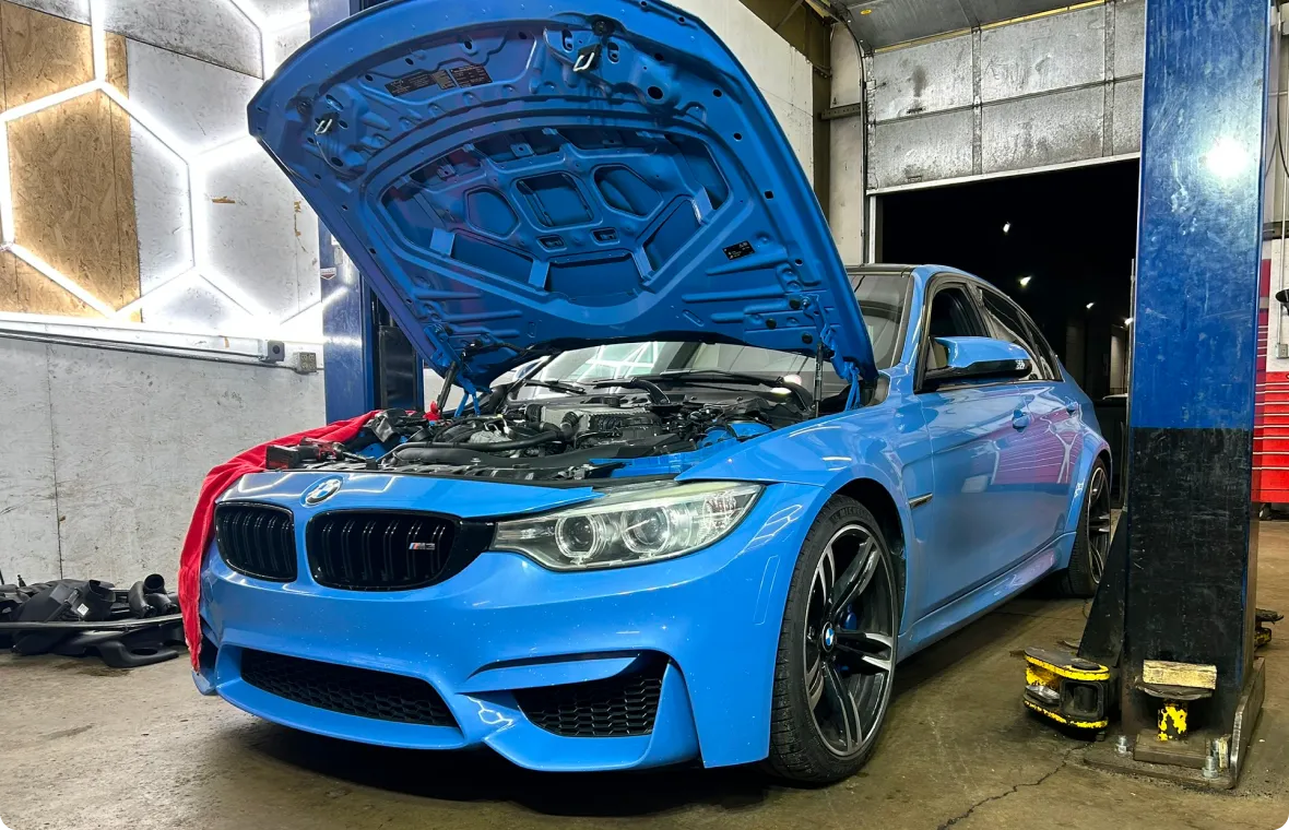 A blue BMW sedan with its hood open is parked in a garage, positioned next to a hydraulic lift. Various car parts and tools are visible around the vehicle, indicating it is being serviced or repaired.