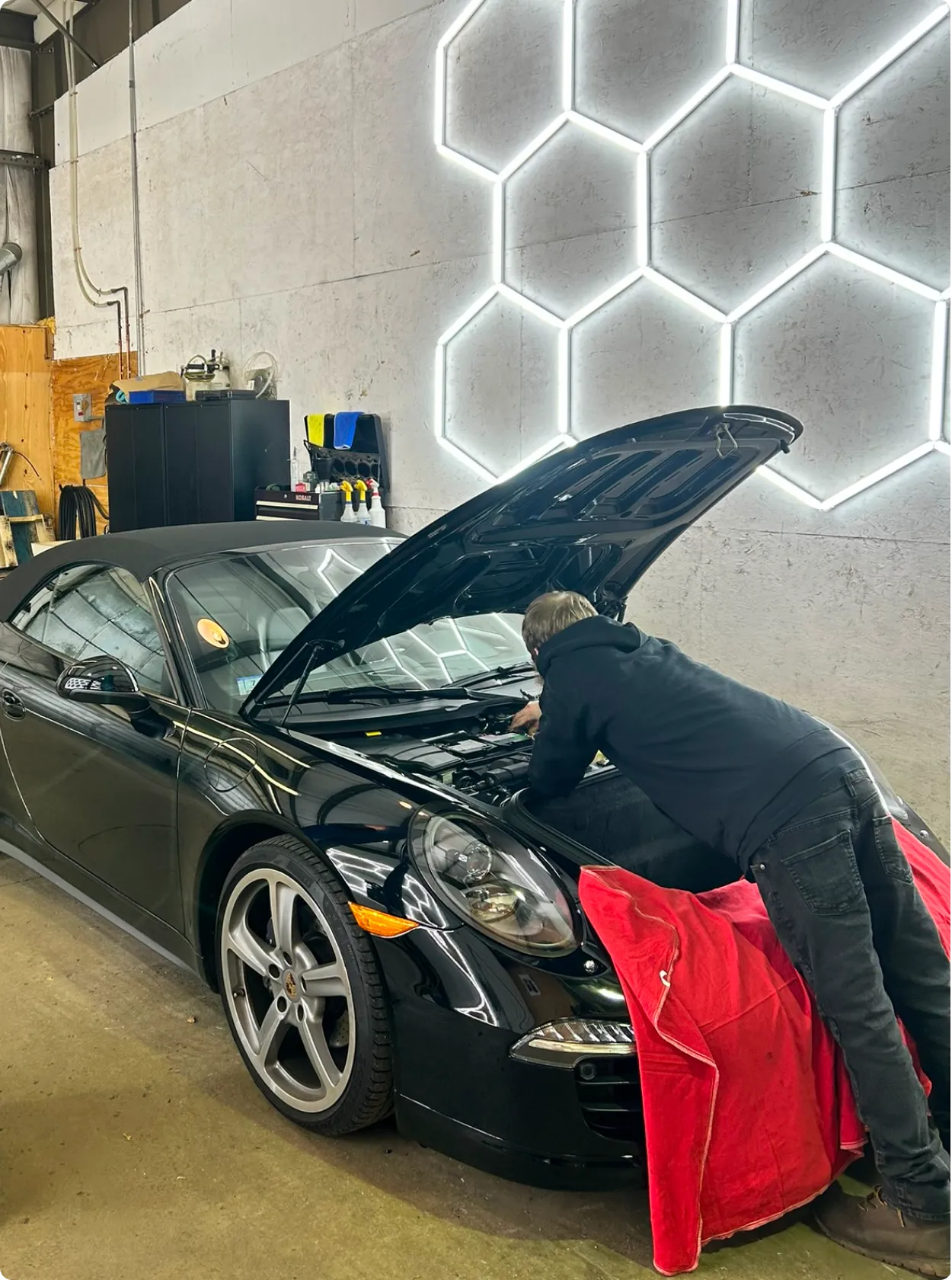 A person in a black hoodie works under the hood of a black convertible car in a garage with a geometric hexagonal light pattern on the wall. A red cloth is draped over the car’s fender.
