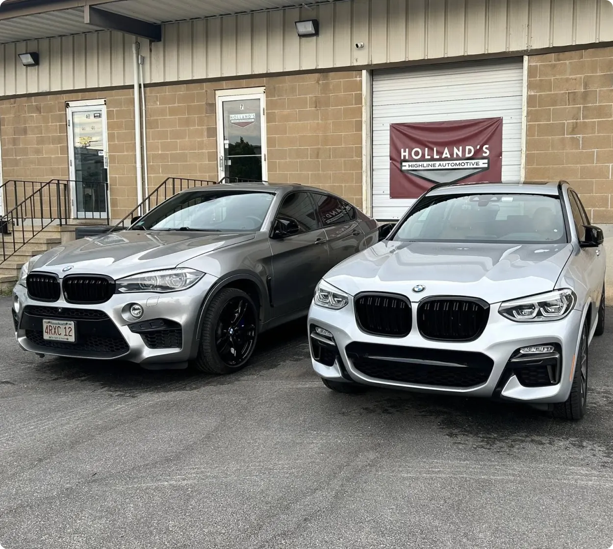 Careers, auto repair in Bridgewater, MA at Holland’s Highline Automotive. Two silver BMW SUVs are parked side by side in front of a building with a brown brick wall and a white garage door featuring a sign that reads Hollands Highline Automotive.