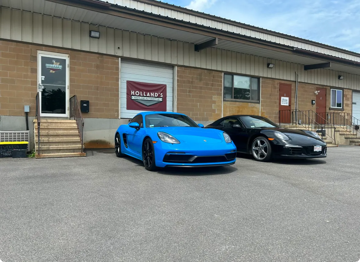 Two sports cars, one bright blue and one black, are parked outside a commercial building with a Hollands sign above the door. The building has tan brick walls, steps, and several doors and windows.