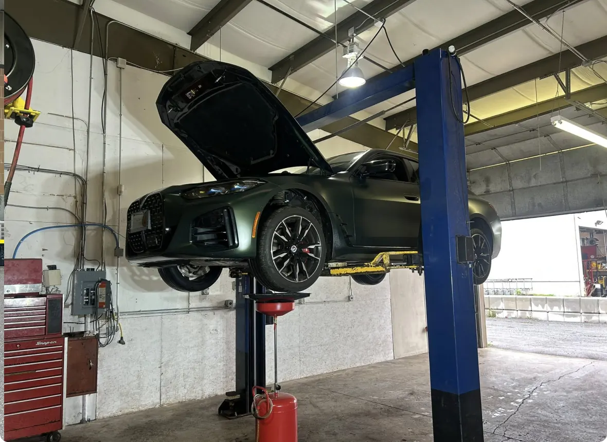 A dark green car with its hood open is lifted on a hydraulic car lift inside an auto repair shop. Tools, equipment, and a red tool chest are visible nearby under fluorescent lighting.