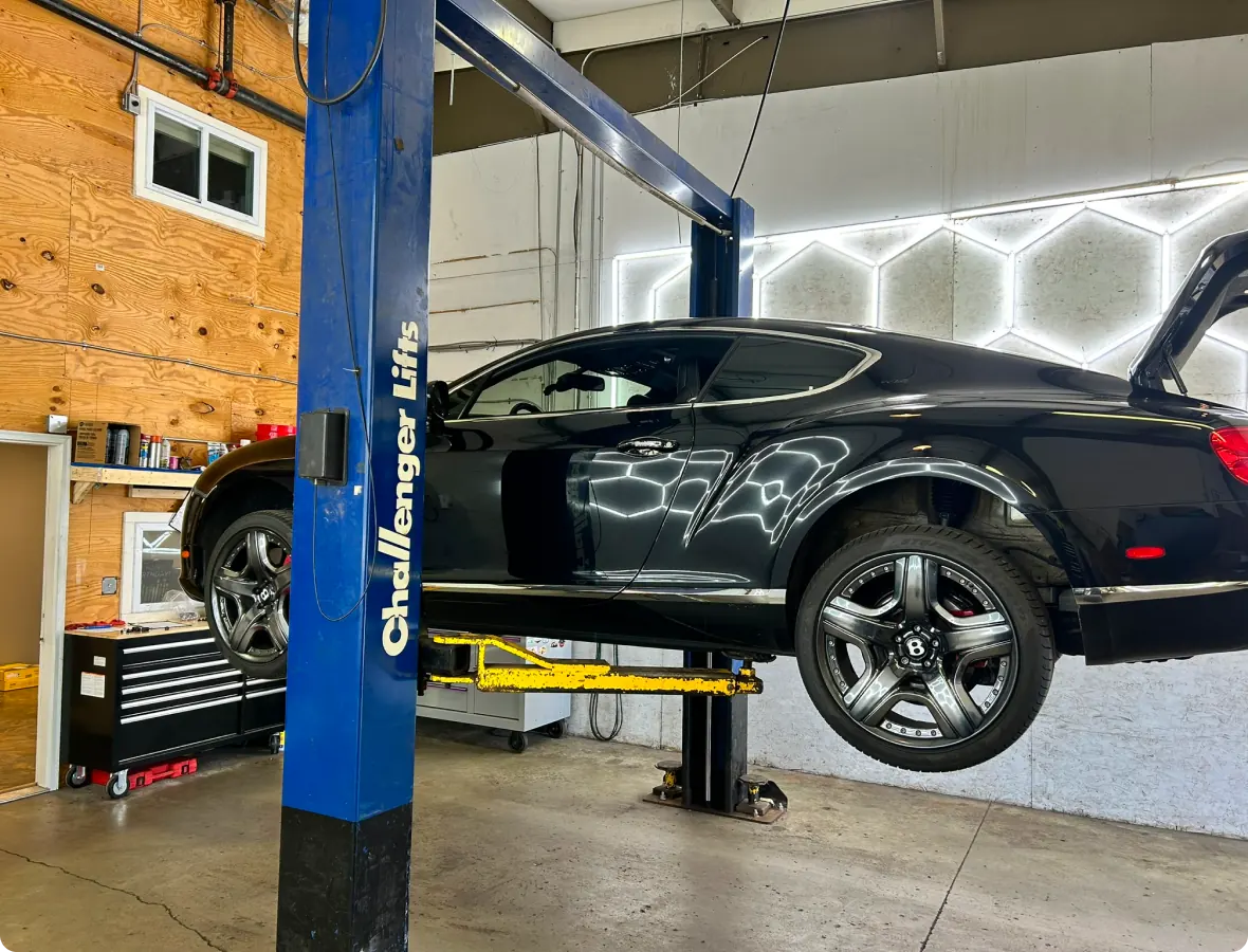 European auto repair in Bridgewater, MA at Holland’s Highline Automotive. A black luxury car is lifted on a blue hydraulic car lift inside a garage. The cars trunk is open, and tools and equipment are visible along the wall and on a workbench in the background.