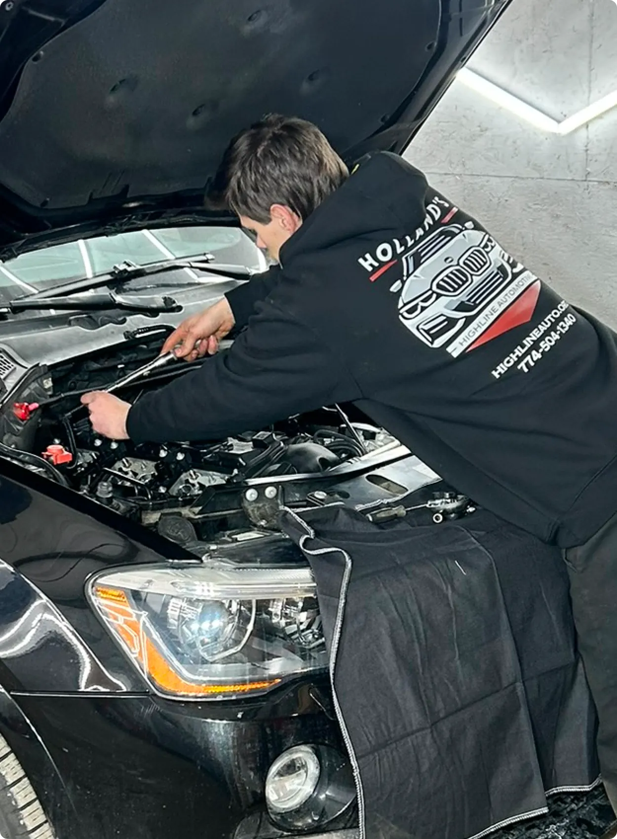A mechanic wearing a black hoodie with a car graphic works on a car engine with the hood open in a well-lit garage.