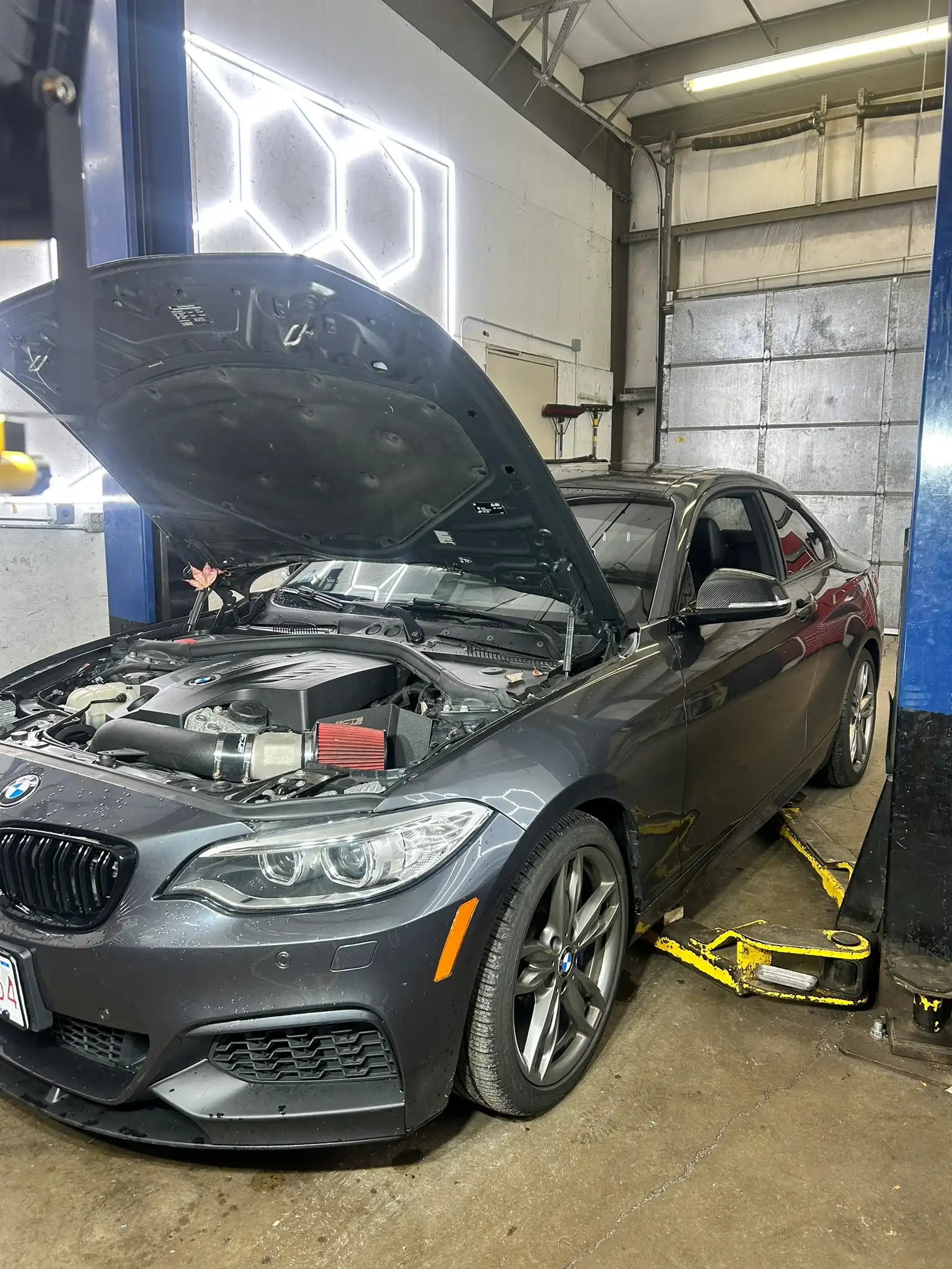 Preventative maintenance in Bridgewater, MA at Holland’s Highline Automotive. A gray BMW car with its hood open is parked inside Holland's Highline Automotive garage in MA on a hydraulic lift, one wheel removed, as technicians perform preventative maintenance Bridgewater locals trust. Garage equipment surrounds the vehicle.