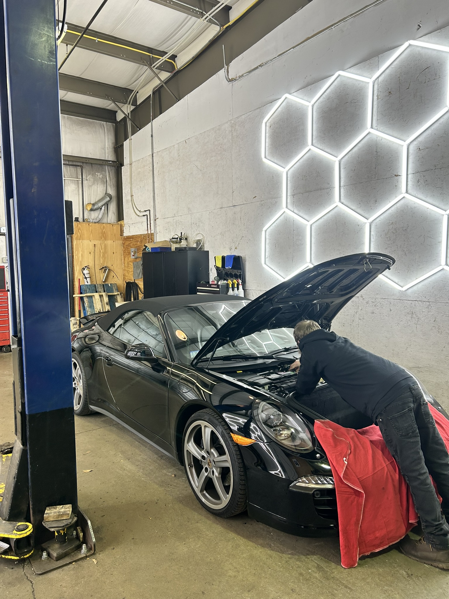 A person works under the hood of a black sports car inside Holland's Highline Automotive in MA, an auto repair shop with industrial lighting and a honeycomb-shaped neon wall light—where diagnostics Bridgewater trusts are performed.
