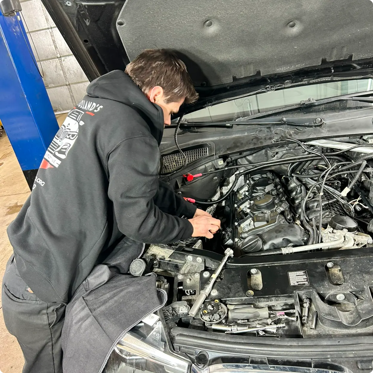 A mechanic in a black hoodie works under the hood of a car, using tools to repair the engine inside an auto repair shop.