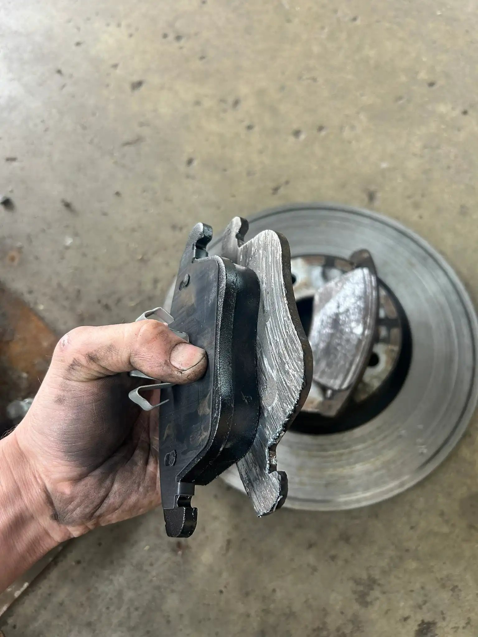 A hand holds two brake pads—one new, one worn—showcasing the importance of preventative maintenance in Bridgewater, MA, with a brake rotor behind. Trust Holland's Highline Automotive for expert care.