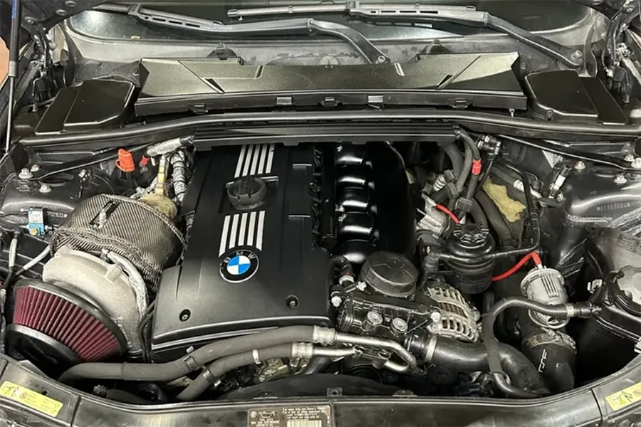 A BMW engine bay featuring a black engine cover with the BMW logo and stripes, visible air intake with cone filter, and various engine components and wiring surrounding the engine block.