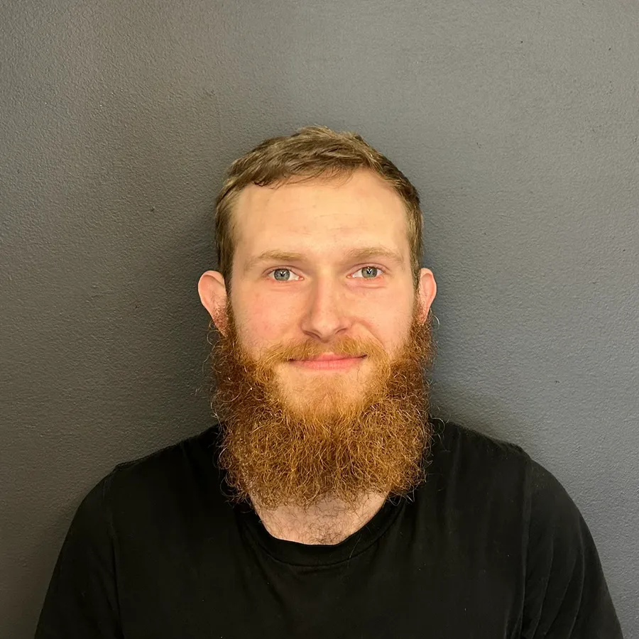 A man with a full reddish-brown beard and short hair is smiling softly at the camera. Wearing a plain black T-shirt, he appears ready to join the team specializing in European auto repair in Bridgewater, posed before a dark gray wall.
