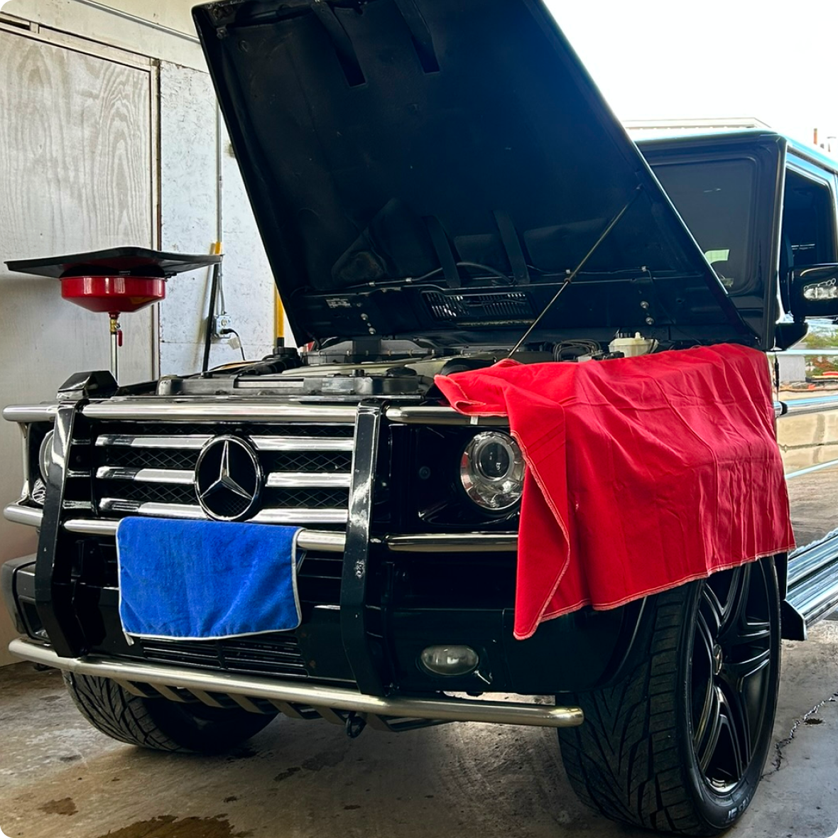 Mercedes repair in Bridgewater, MA at Holland’s Highline Automotive. A black Mercedes-Benz SUV with its hood open is parked in a garage at Holland's Highline Automotive, Bridgewater, MA. A red cloth is draped over the right fender and a blue cloth covers the grille as diagnostics are being performed.