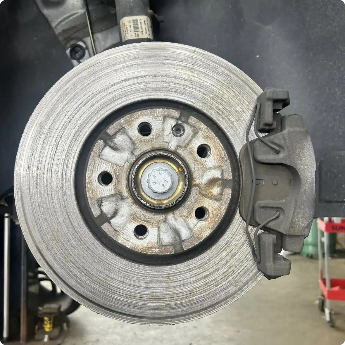 Close-up view of a car's brake disc and caliper at Holland's Highline Automotive. The rotor shows wear marks, and the brake caliper is mounted to the right—wheel removed for maintenance such as brake fluid service or brake repair Bridgewater.