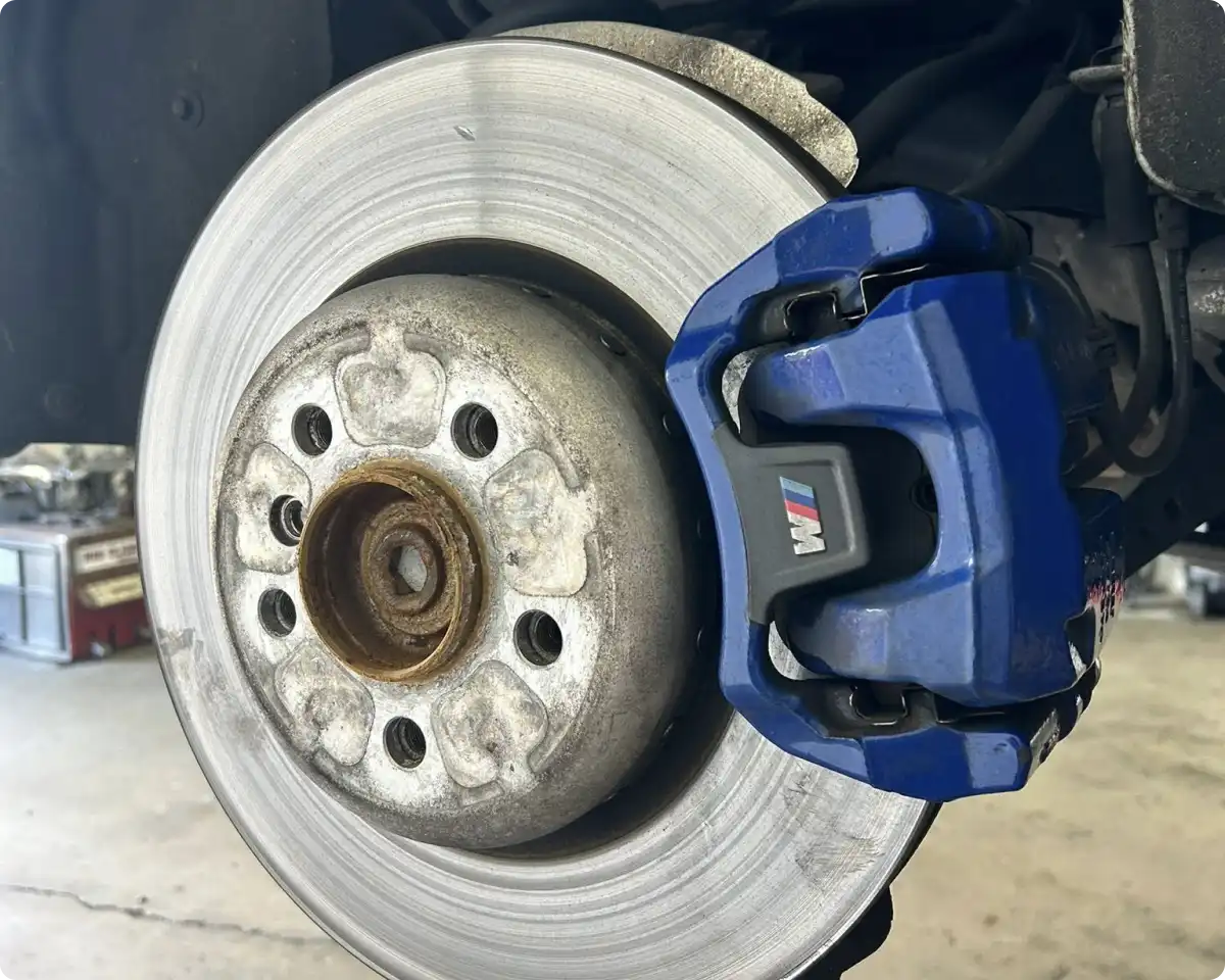 Brake repair in Bridgewater, MA at Holland’s Highline Automotive. Close-up of a car’s front brake disc and a blue caliper with the BMW M logo, showing rust on the hub, inside Holland's Highline Automotive workshop—perfect for those seeking expert brake pad replacement or brake repair Bridgewater.