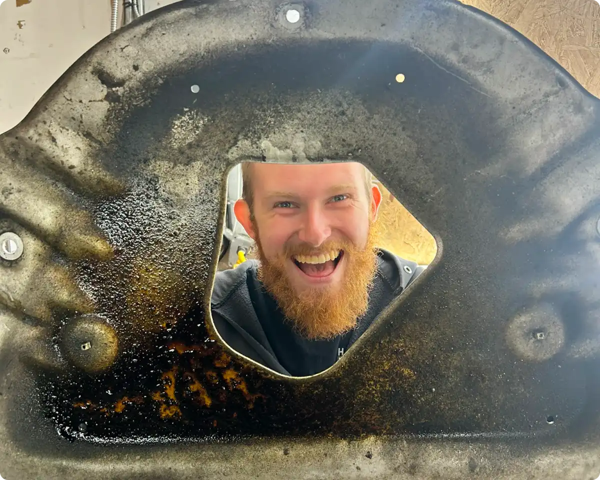 Vehicle diagnostics in Bridgewater, MA at Holland’s Highline Automotive. A bearded man with a big smile looks through a roughly diamond-shaped hole in a large, greasy, metallic object at Holland's Highline Automotive, MA—your go-to spot for diagnostics in Bridgewater.
