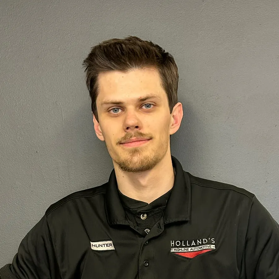 A young man with short brown hair and light facial hair stands in front of a gray wall, wearing a black collared Hollands Discipline Automotive shirt with a name tag that says Hunter, ready to assist the team with expert European auto repair.