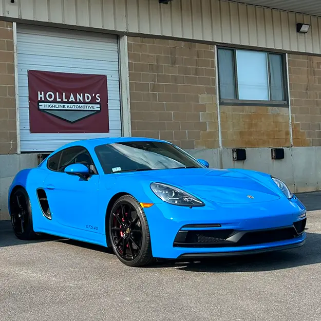 A bright blue Porsche sports car with black rims is parked in front of Holland's Highline Automotive, a top spot for Porsche repair in Bridgewater, MA, and a building with a closed white garage door.