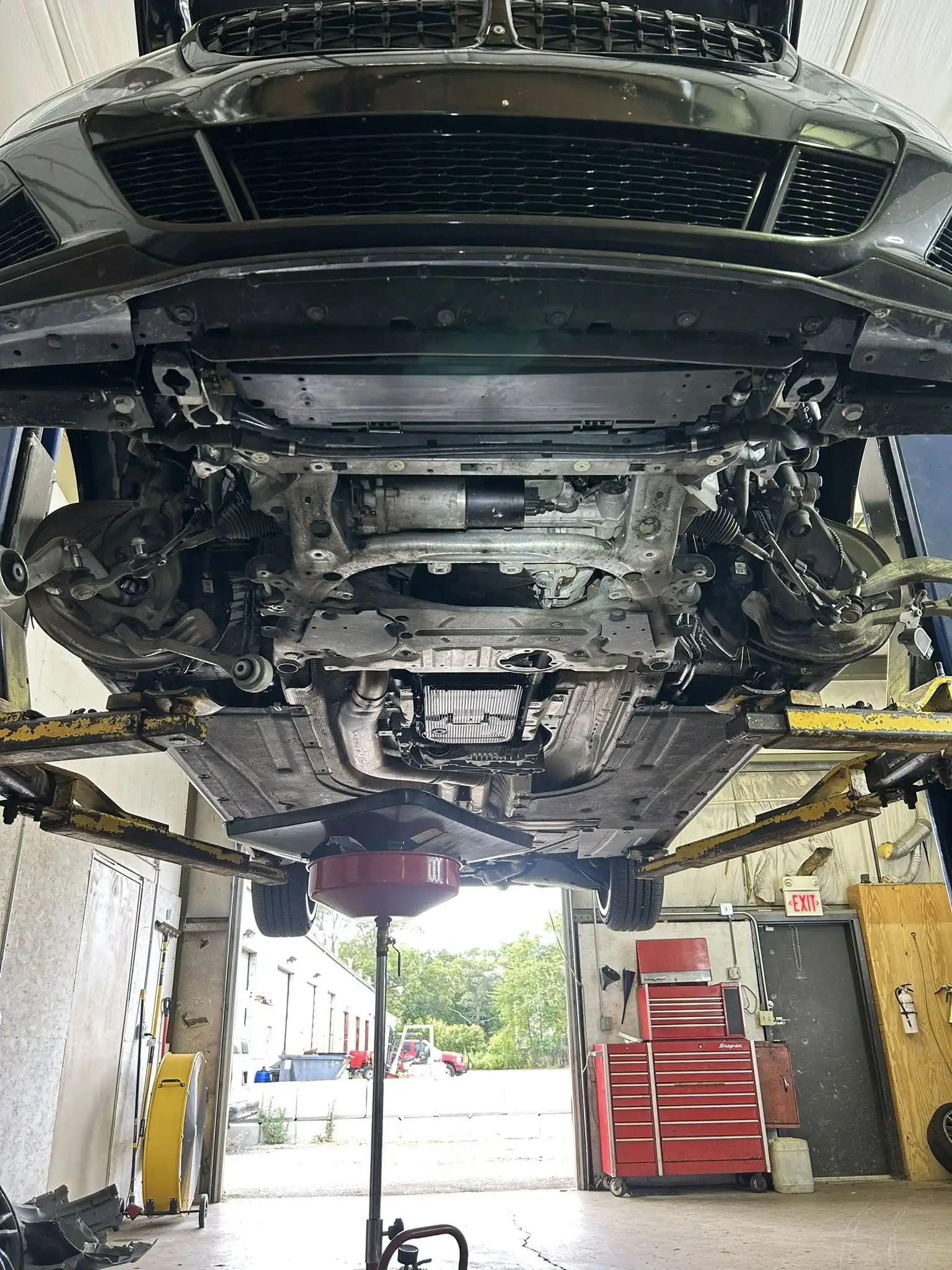 Oil change in Bridgewater, MA at Holland’s Highline Automotive. A car raised on a hydraulic lift at Holland's Highline Automotive in MA reveals its undercarriage. Tools, red tool chests, and equipment fill the shop—perfect for an oil change in Bridgewater with garage doors open in the background.