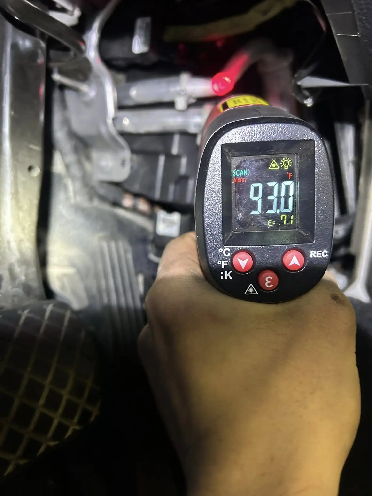 A hand holds an infrared thermometer aimed at a car part, displaying 93.0°C on the screen—perfect for diagnosing issues during heater repair. The car’s pedals and interior parts are visible in the background.