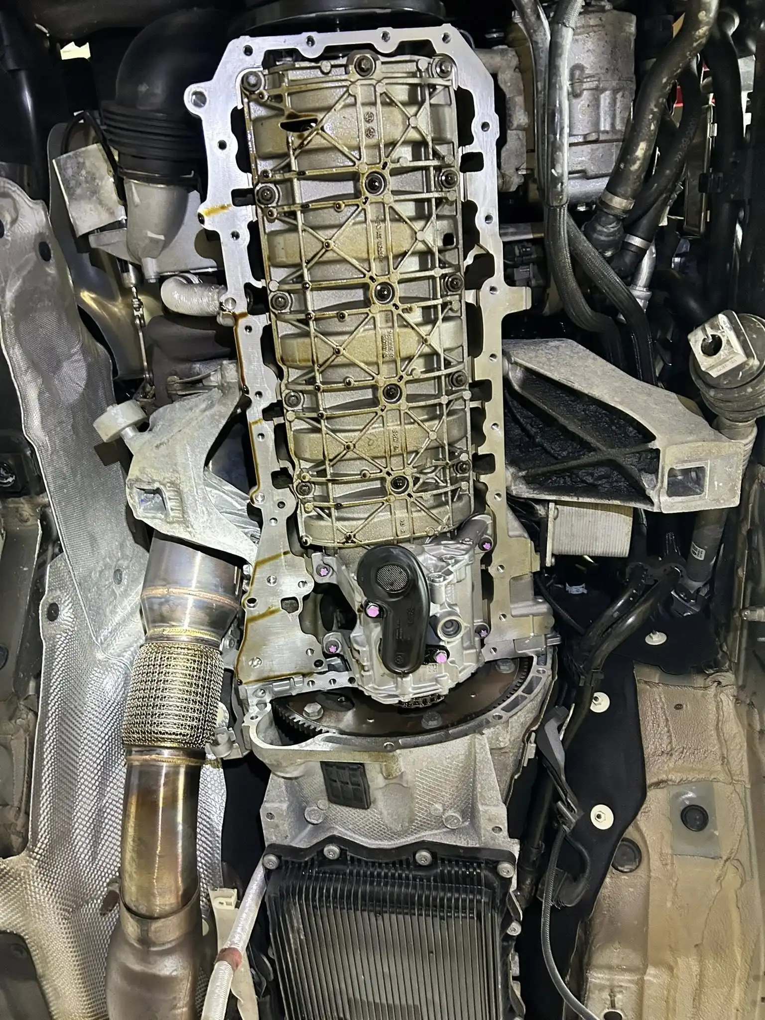 Close-up view of a car engine’s underside during an oil change Bridgewater, MA, with the oil pan removed to reveal internal components, metal parts, visible wiring, and exhaust pipe surrounded by various mechanical parts.