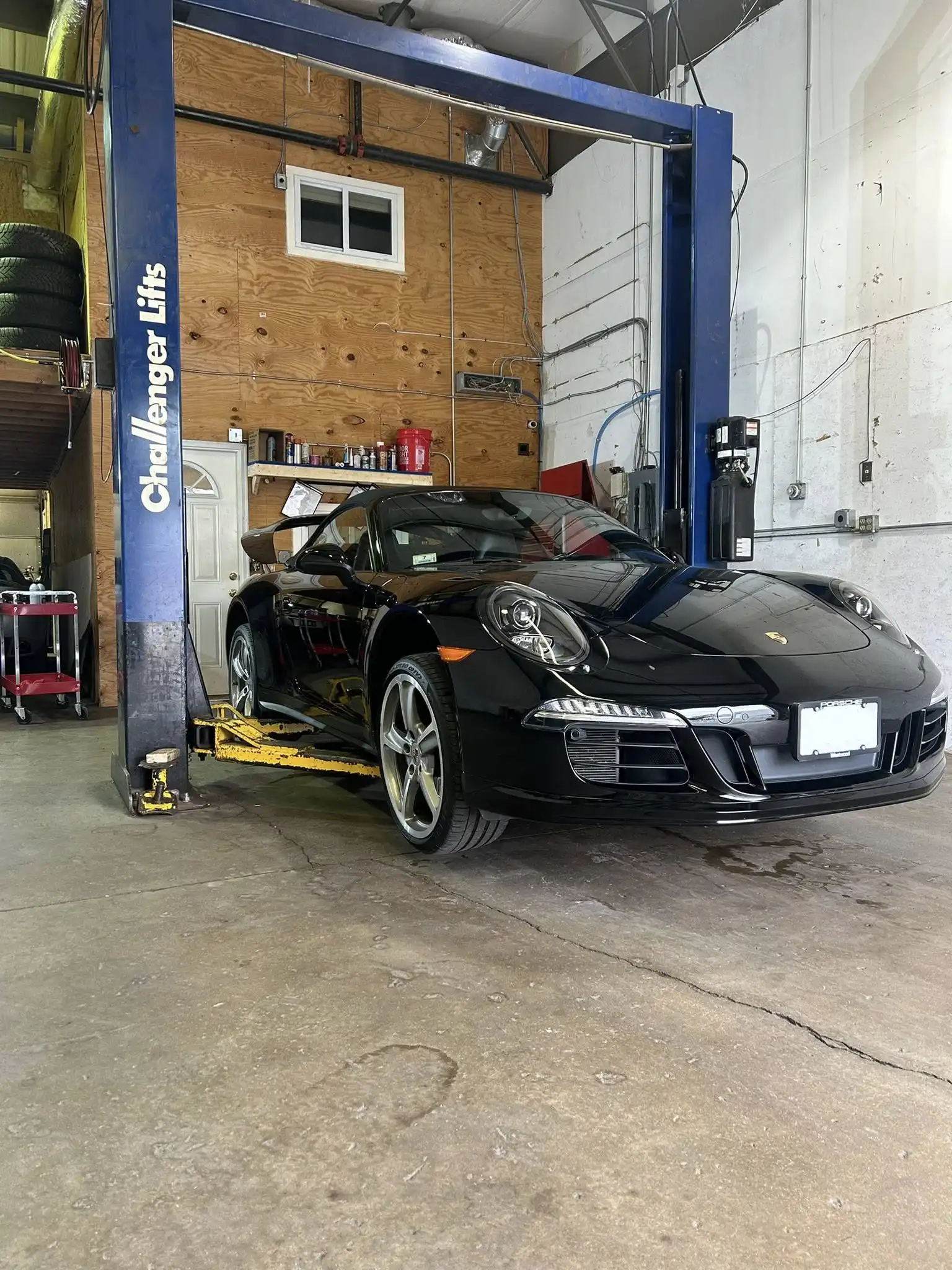 A black Porsche sports car is parked under a blue vehicle lift inside Holland's Highline Automotive, an auto repair shop in MA specializing in auto a/c repair Bridgewater, with wooden and white walls, shelves with tools, and stacked tires visible in the background.