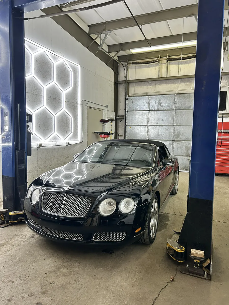 A black Bentley car is parked inside an auto repair shop, positioned under a vehicle lift for Bentley repair. The room features a metal garage door, a honeycomb light fixture on the wall, and a red tool chest in the corner.