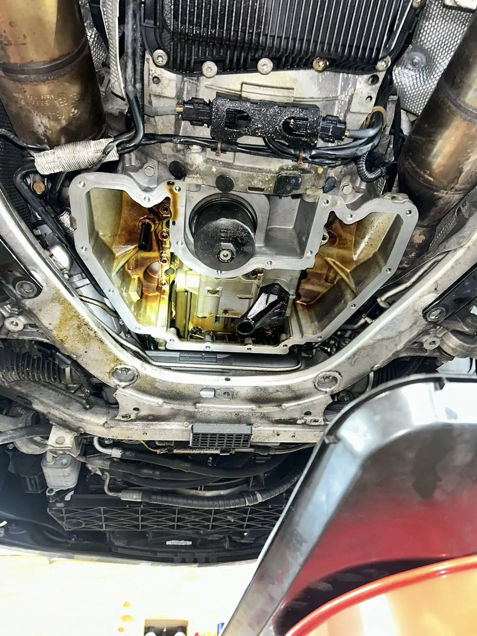 View of the underside of a car at Holland's Highline Automotive, MA, with the oil pan removed to expose engine components. A mechanic’s oil drain pan is positioned below to collect fluids during a synthetic oil change.
