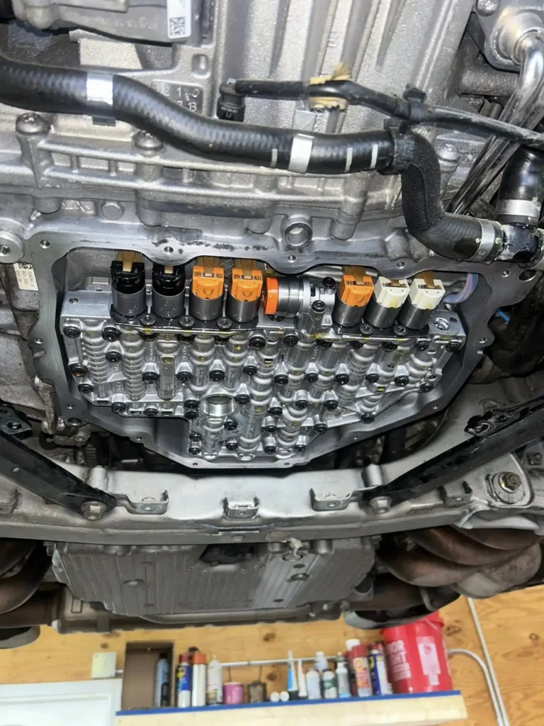 Close-up view of the underside of a car showing the exposed automatic transmission valve body with several solenoids in different colors, surrounded by metal components and wiring.