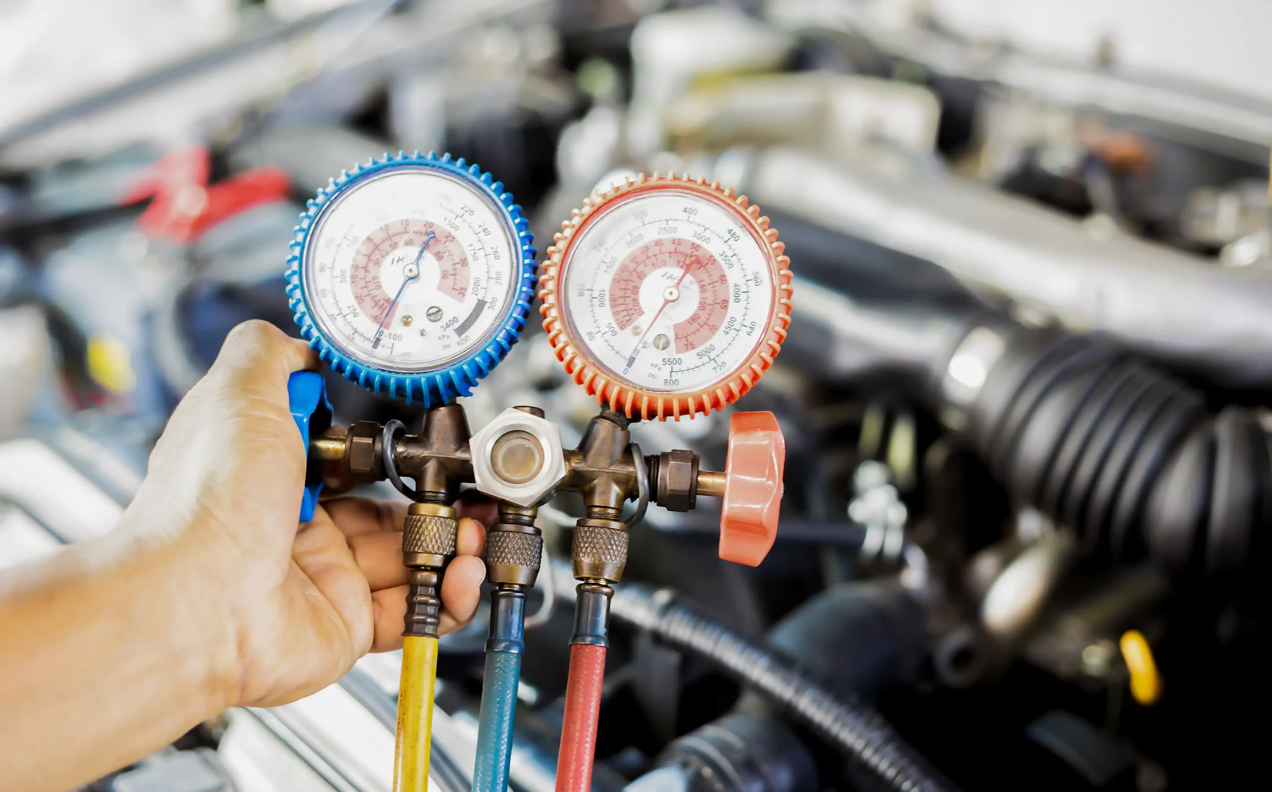 Auto a/c & heater repair in Bridgewater, MA at Holland’s Highline Automotive. A person holds a manifold gauge set, with blue and red dials, used for checking air conditioning or refrigeration systems in front of an open car engine—essential for auto a/c repair Bridgewater and heater repair in MA.