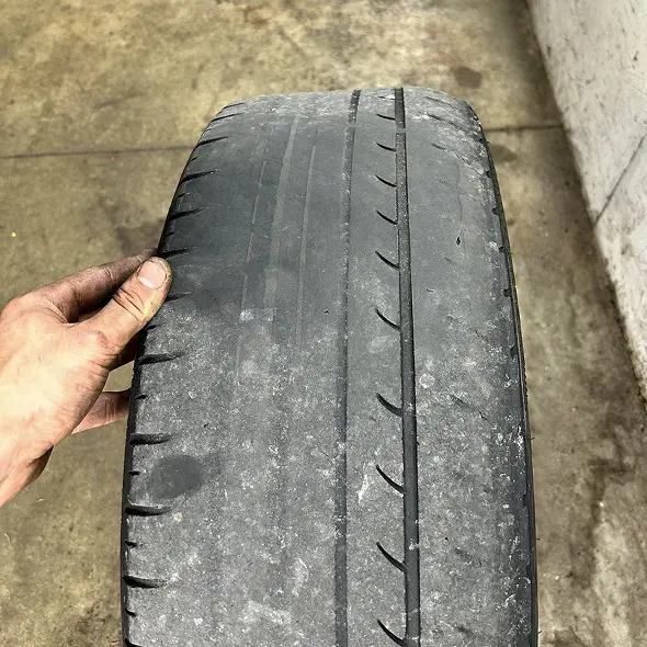 A hand holds a worn car tire with severely faded tread and visible balding, indicating heavy use and the need for replacement. For expert tire service and tire rotation, visit Holland's Highline Automotive. Concrete background completes the scene.