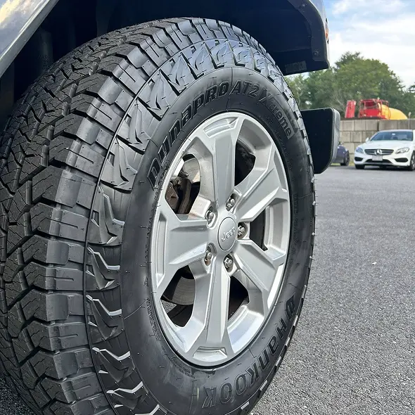 Close-up of a Jeep vehicle’s front tire with Dynapro AT2 and Hankook branding, parked on asphalt near other vehicles and trees—captured at Holland's Highline Automotive, your trusted spot for tire repair in Bridgewater, MA.