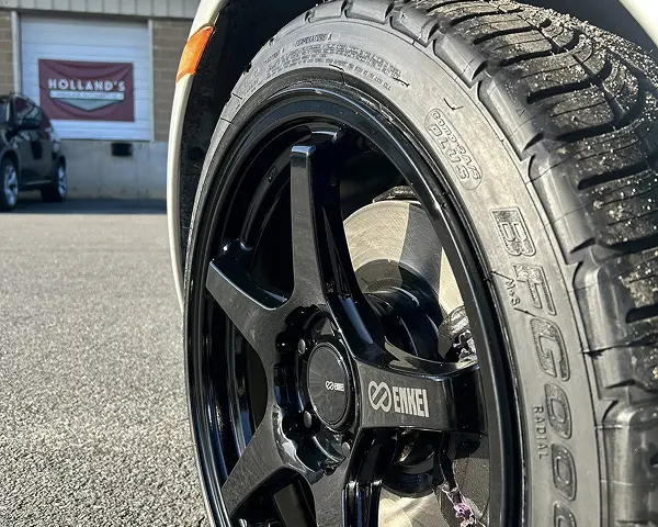 Tire repair and services in Bridgewater, MA at Holland’s Highline Automotive. Close-up of a black Enkei car wheel and tire parked on asphalt, with a building and a Holland’s Highline Automotive sign visible in the blurred background, hinting at expert tire service nearby.