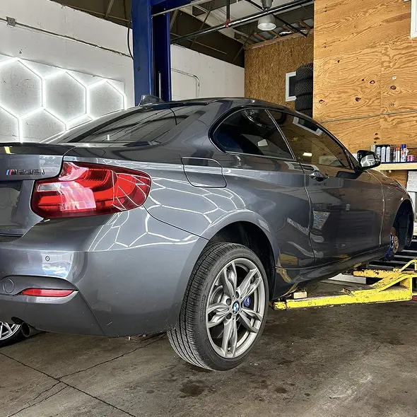 A gray BMW coupe is lifted on a yellow hydraulic car lift at Holland's Highline Automotive in MA, specializing in suspension repair Bridgewater, with tools and equipment visible in the background.