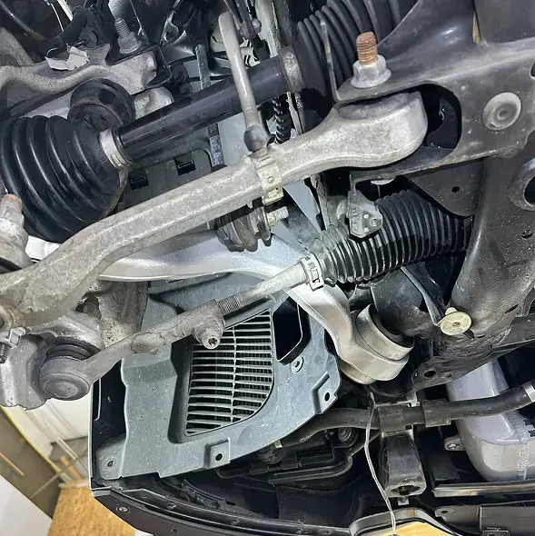 Close-up view of a car’s front suspension and steering components, including control arms, tie rod, axle, and protective boots, taken from underneath the vehicle during suspension repair at Holland's Highline Automotive in Bridgewater, MA.