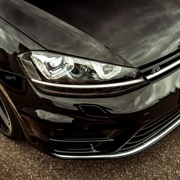 Close-up of the front left side of a shiny black car, showing the headlight, grille, and hood with cloudy sky reflections on its surface. Parked on a paved road near Holland's Highline Automotive, your MA destination for Volkswagen repair Bridgewater.