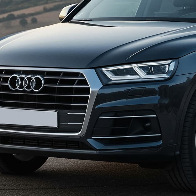 Close-up of the front of a dark-colored Audi SUV, featuring the grille with the Audi logo, sleek headlights, and part of the hood and wheel—captured outside Holland's Highline Automotive, your trusted spot for Audi repair in Bridgewater, MA.