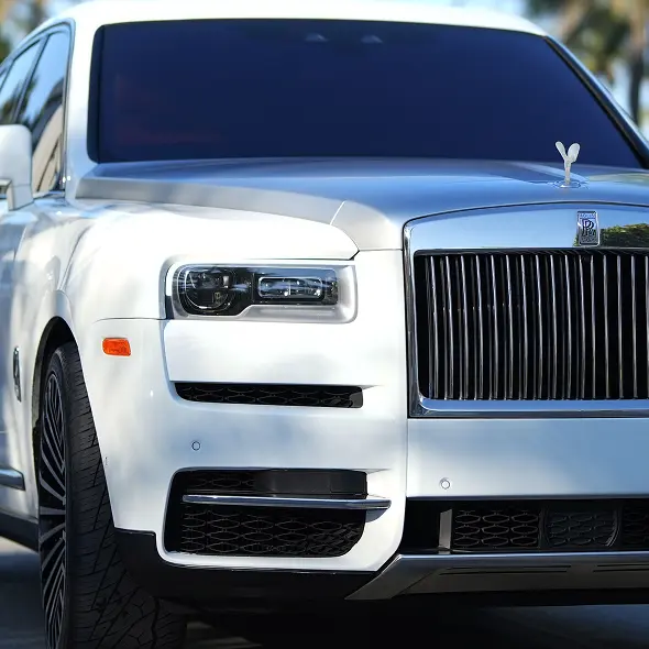 Close-up view of a white Rolls-Royce SUV at Holland's Highline Automotive, Bridgewater, MA, showcasing its iconic grille, Spirit of Ecstasy ornament, and modern headlights. The luxury car is parked outdoors with blurred greenery in the background.