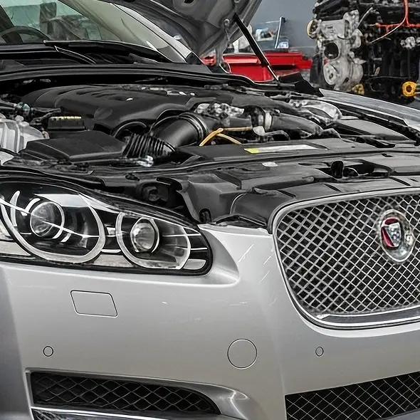 A close-up of a silver Jaguar with its hood open, revealing the engine. The front grille and headlights are visible, with another engine in the background at Holland's Highline Automotive, your Jaguar repair experts in Bridgewater, MA.