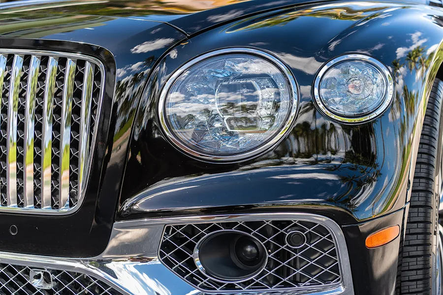 Close-up of the front end of a shiny black luxury car, possibly in for Bentley repair, showing its grille, two circular headlights, and a portion of the tire. Reflections of trees and sky are visible on the car’s polished surface.
