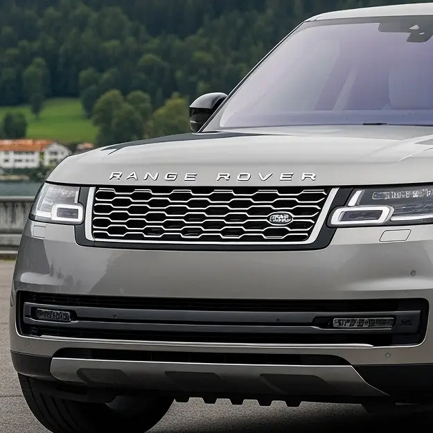 Close-up of the front of a silver Range Rover SUV at Holland's Highline Automotive in MA, highlighting its grille, headlights, and part of the hood; greenery and buildings blur in the background. Land Rover repair Bridgewater specialists.