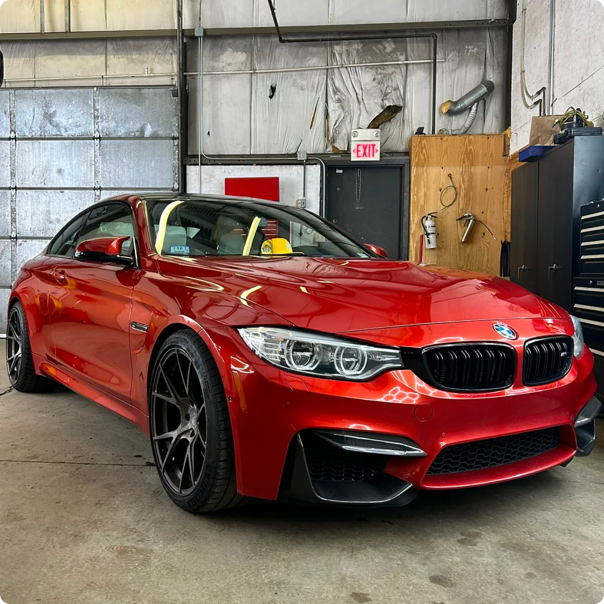 A shiny red BMW coupe with black wheels is parked inside a garage with concrete floors, metal walls, and an exit sign—an ideal setting for top-notch BMW repair & service.