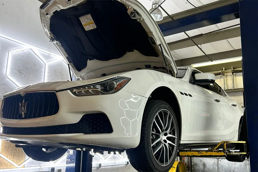 A white Maserati sedan with its hood open is elevated on a hydraulic lift inside an auto repair shop, showing the front grille, left headlight, and left front tire.