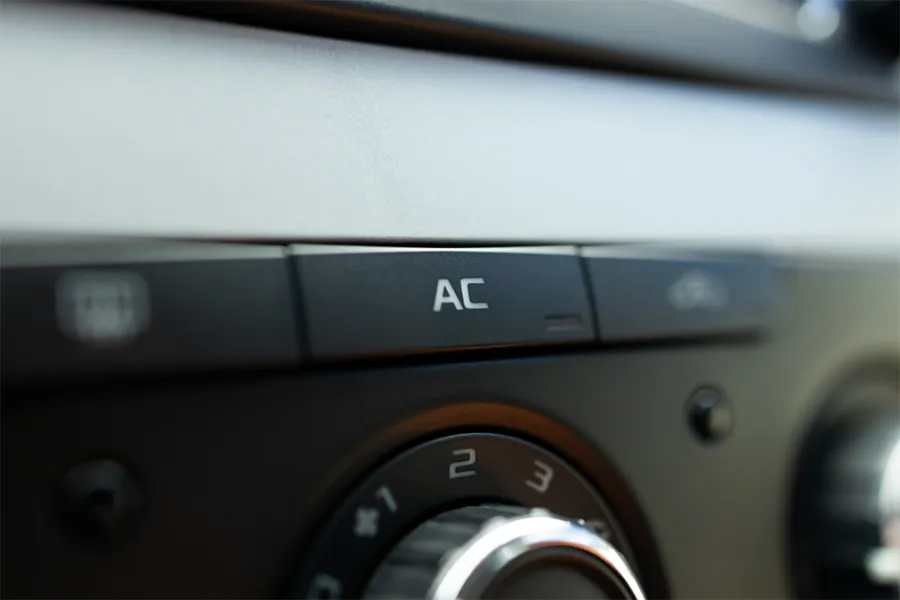 Close-up of a car dashboard showing the AC button and part of the temperature control dial. The image focuses on the air conditioning controls.