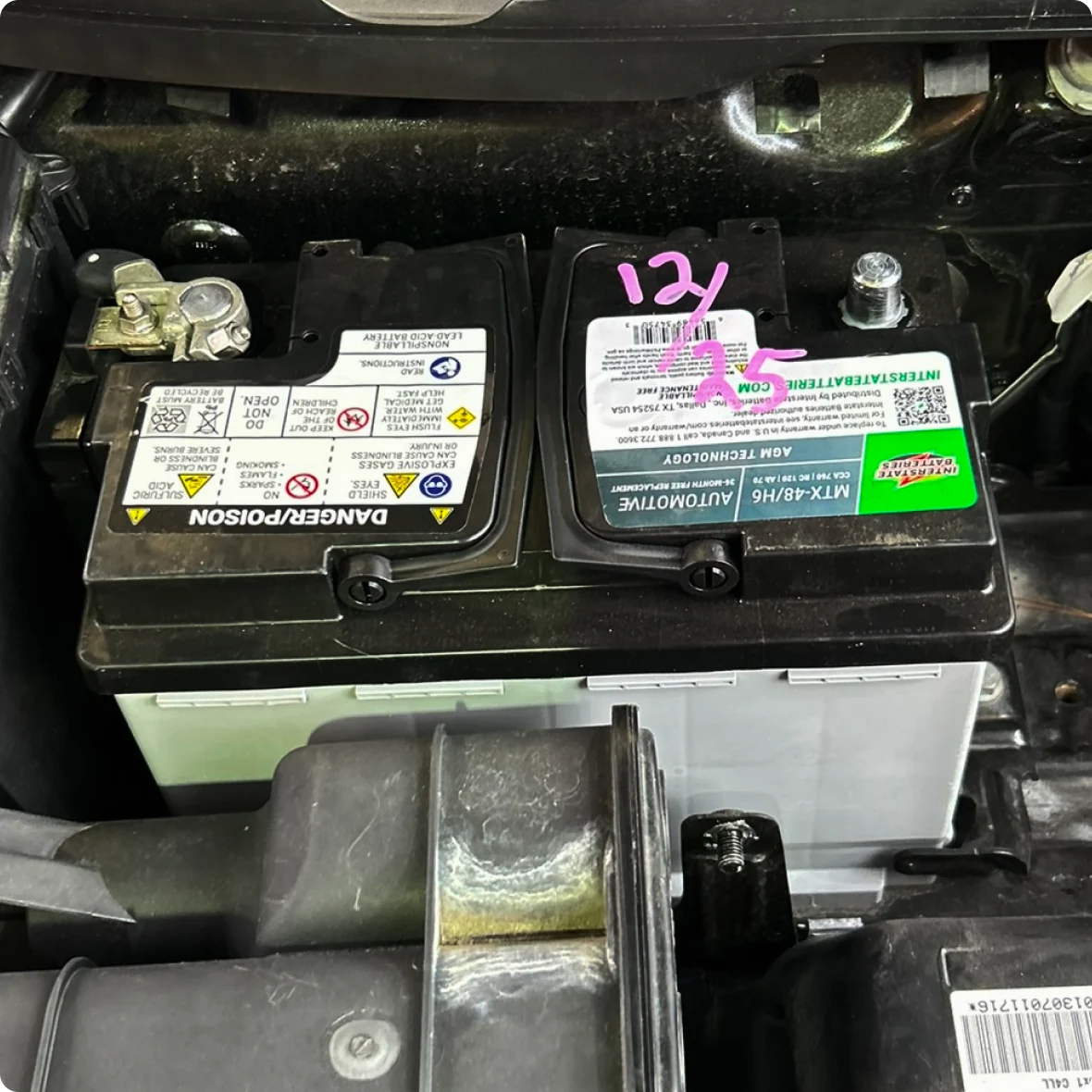 A close-up of two car batteries side by side under a hood; the right battery—marked “12V” and “75” in pink—features warning labels, ideal for those seeking auto electrical repair or servicing a European vehicle.