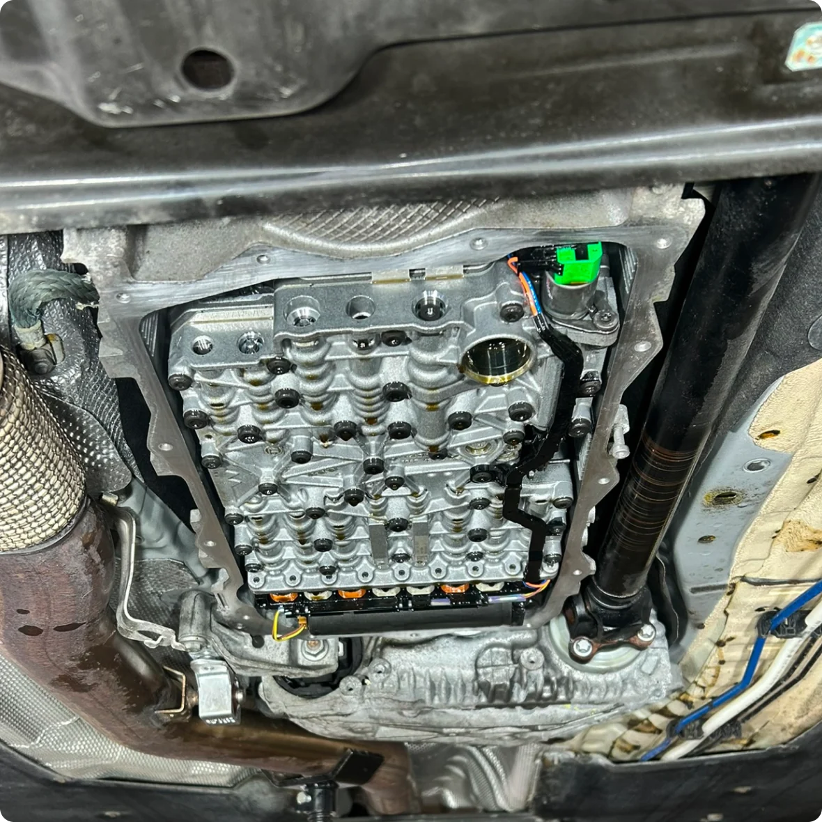 View of a car’s underside showing an exposed automatic transmission valve body with various bolts, connectors, and wiring visible, likely during a transmission service or repair procedure.