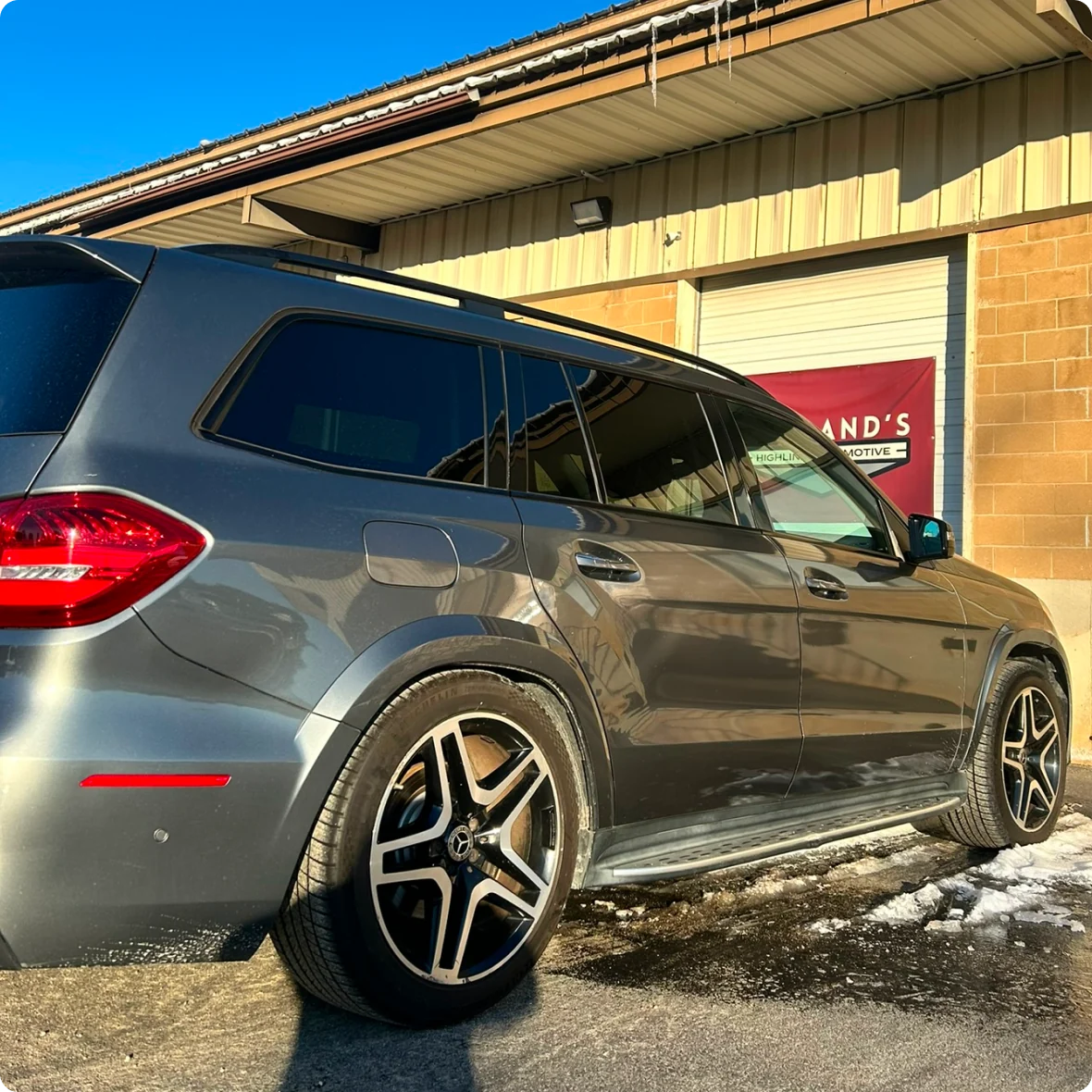 A silver Mercedes-Benz SUV, possibly awaiting mercedes repair in Bridgewater, is parked outside a beige industrial building with a red sign, on a sunny day with patches of melting snow near the car.